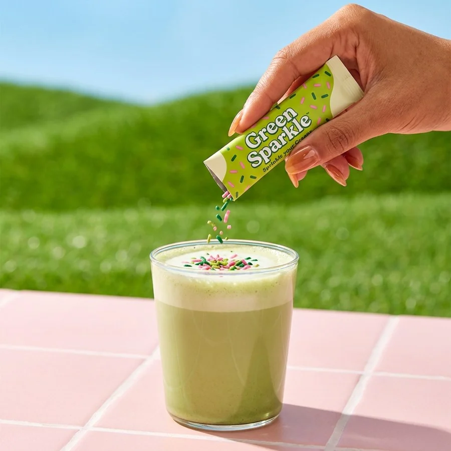 A hand sprinkling a Green Sparkle stick over a matcha latte on a pink tile table